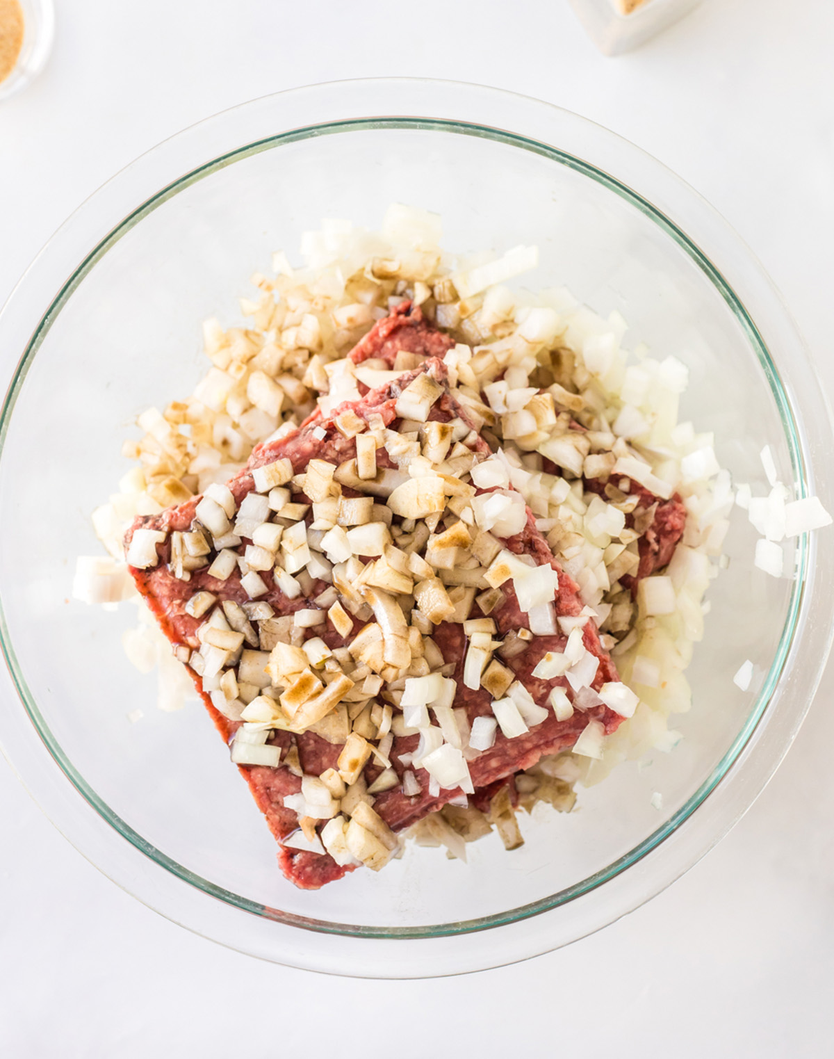 Raw ground beef in a glass bowl with burger ingredients overhead