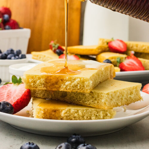 Maple syrup pouring over a stack of fluffy vegan sheet pan pancakes with fresh strawberries and blueberries.