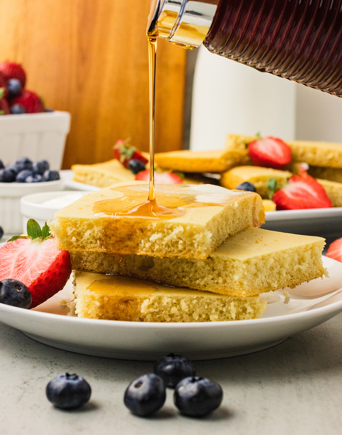 Maple syrup pouring over a stack of fluffy vegan sheet pan pancakes with fresh strawberries and blueberries.