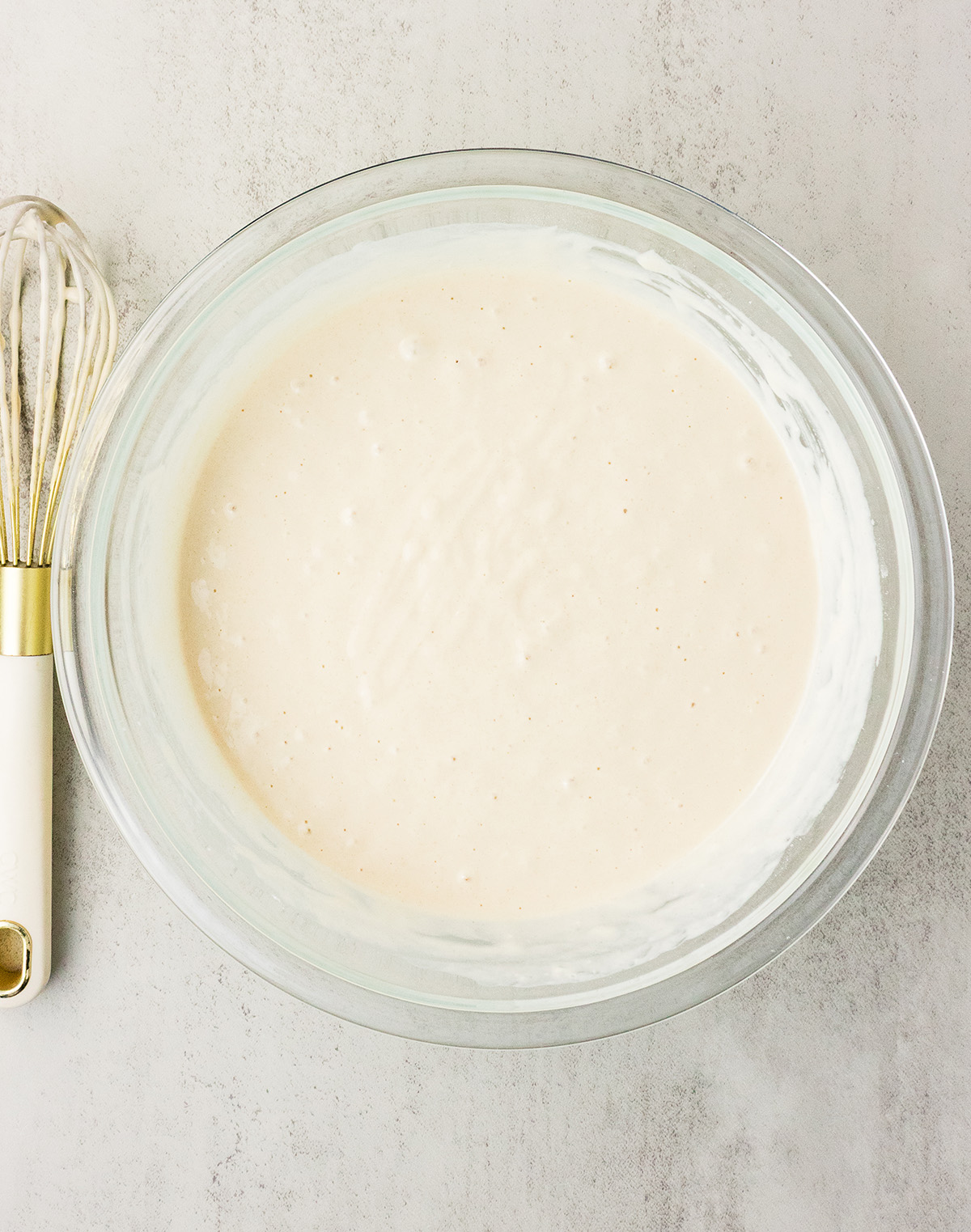 Smooth vegan pancake batter in a glass mixing bowl with a gold and white whisk beside it.